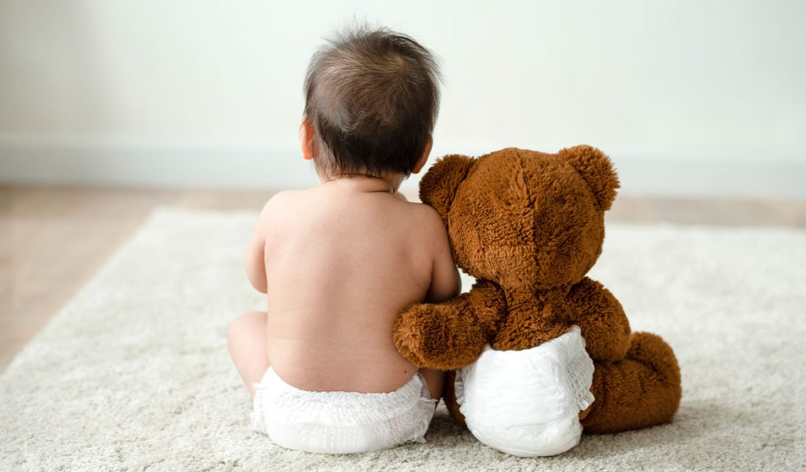 Bebé sentado en el suelo junto a un peluche, ambos con pañal, representando el cuidado de la dermatitis del pañal